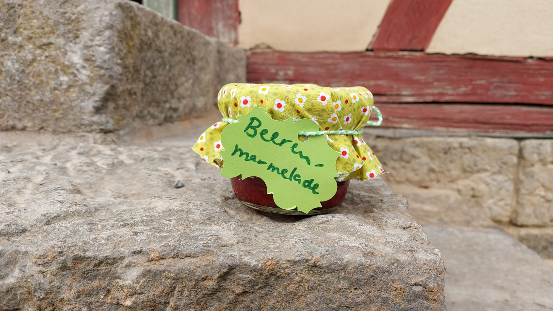 Ein Glas mit grünem Stoffüberzug und Etikett mit der Beschriftung "Beerenmarmelade" steht auf einer Steintreppe vor einer Fachwerkwand