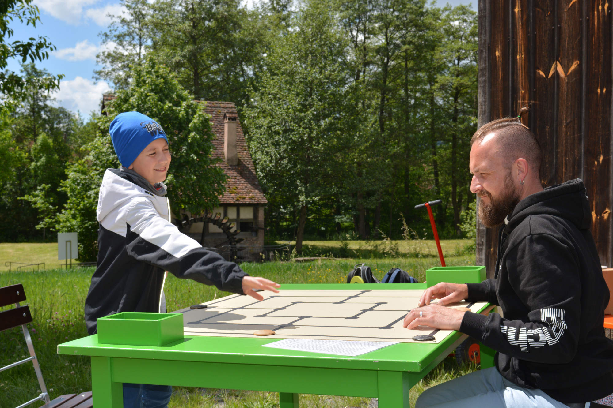 Ein Junge und ein Mann spielen eine Partie M&uuml;hle im Freien, im Hintergrund Feld, Wald und eine Wasserm&uuml;hle