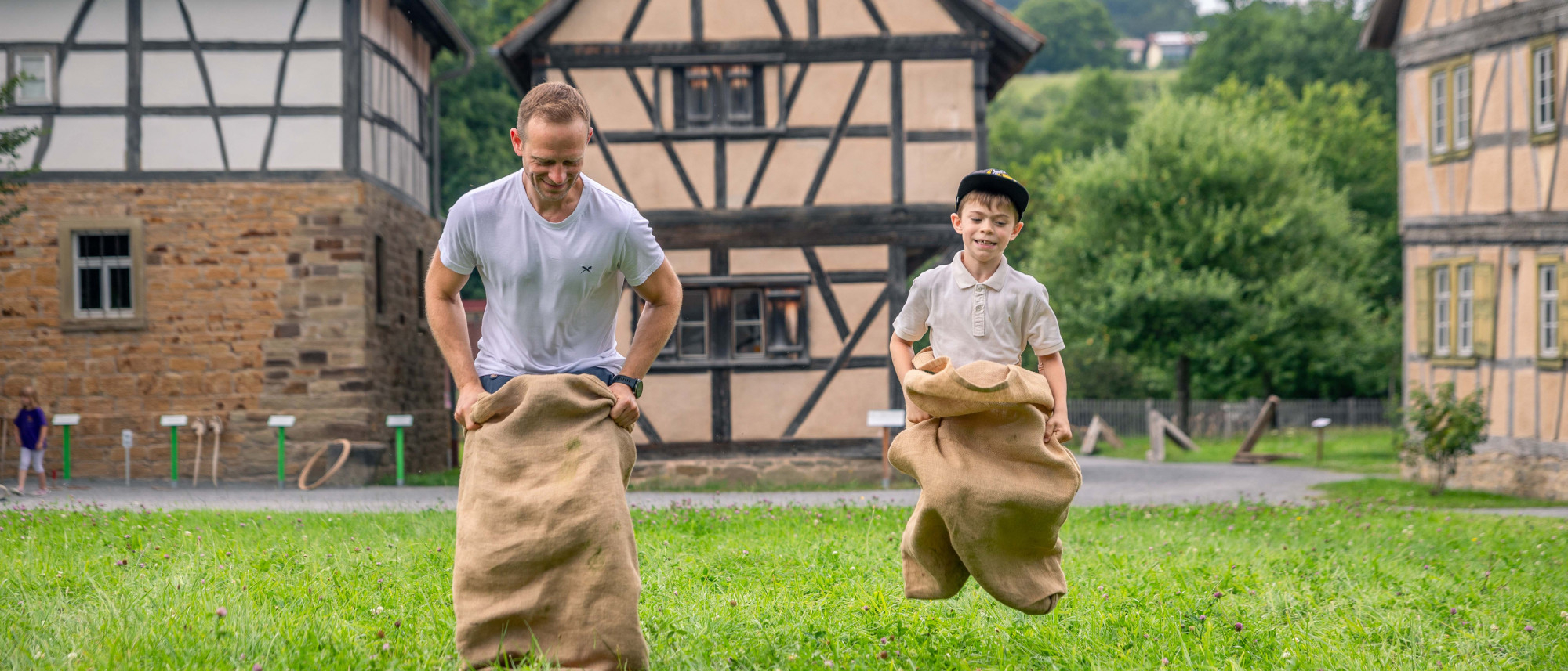 Vater und Sohn machen Sackh&uuml;pfen auf einer Wiese vor Fachwerkgeb&auml;uden