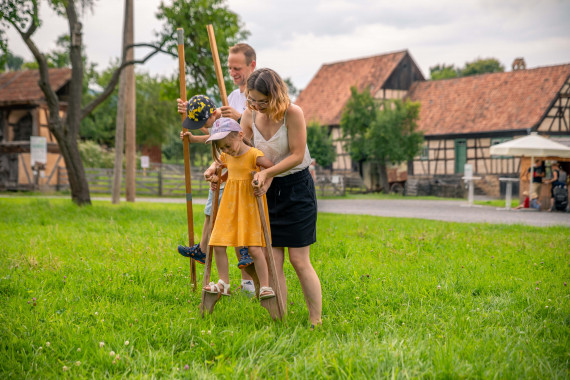 Zwei Kinder &uuml;ben mit ihren Eltern Stelzenlaufen auf einer Wiese, im Hintergrund Fachwerkh&auml;user