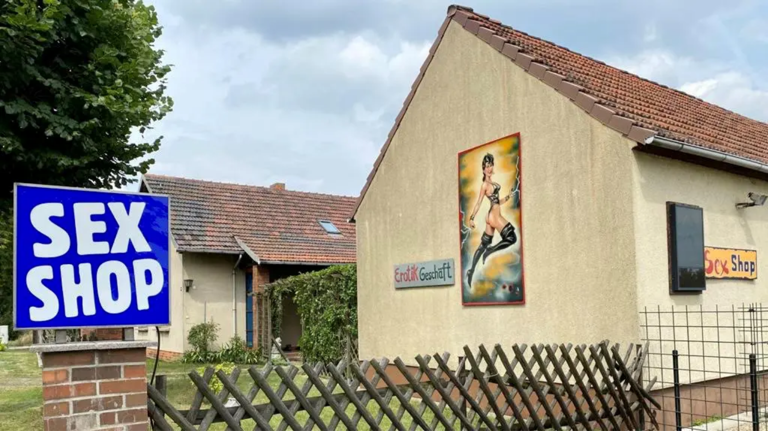 &quot;Sex Shop&quot; Schild neben Gartenzaun, rechts eine Hausfassade mit Werbeplakat und Schild &quot;Erotik Geschäft&quot;