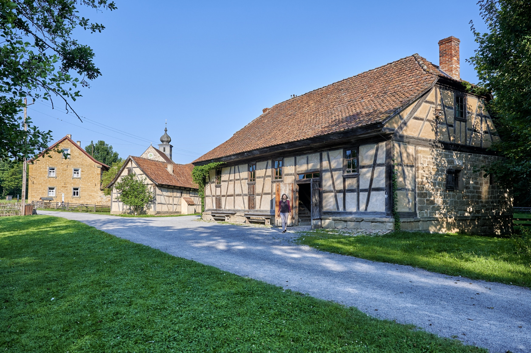 Frau spaziert aus einem Fachwerkgeb&auml;ude, im Vordergrund Schotterweg und Wiese, links und rechts B&auml;ume
