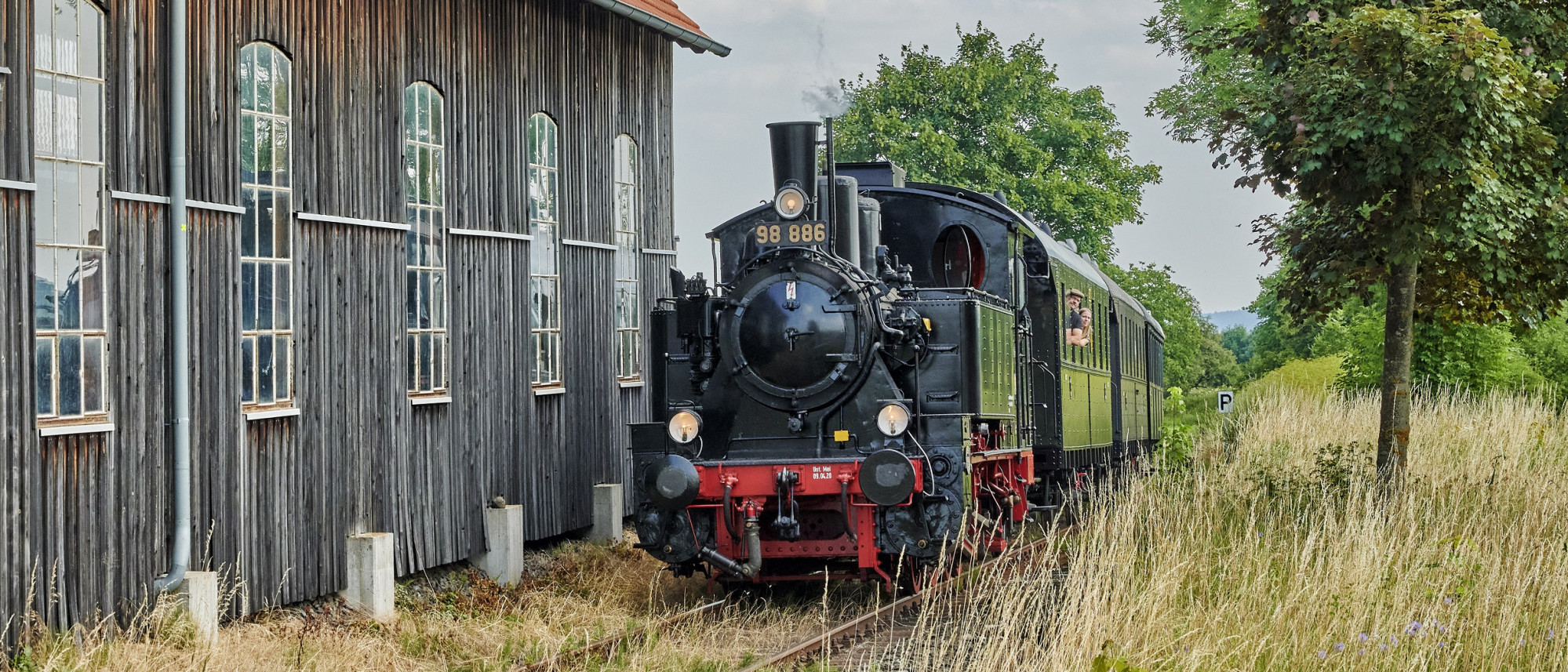 Dampfeisenbahn f&auml;hrt zwischen B&auml;umen und Lokschuppen entlang
