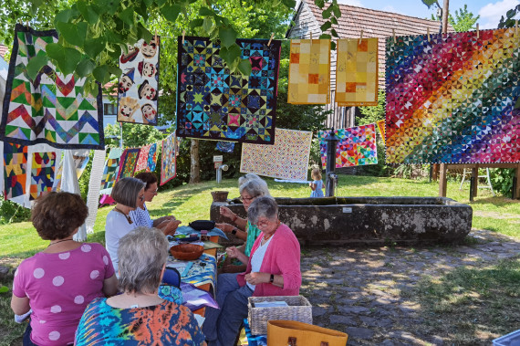 Gruppe &auml;lterer Frauen sitzt an einem Tisch im Freien unter B&auml;umen, im Hintergrund h&auml;ngen bunte Patchwork-Quilts an einer Leine