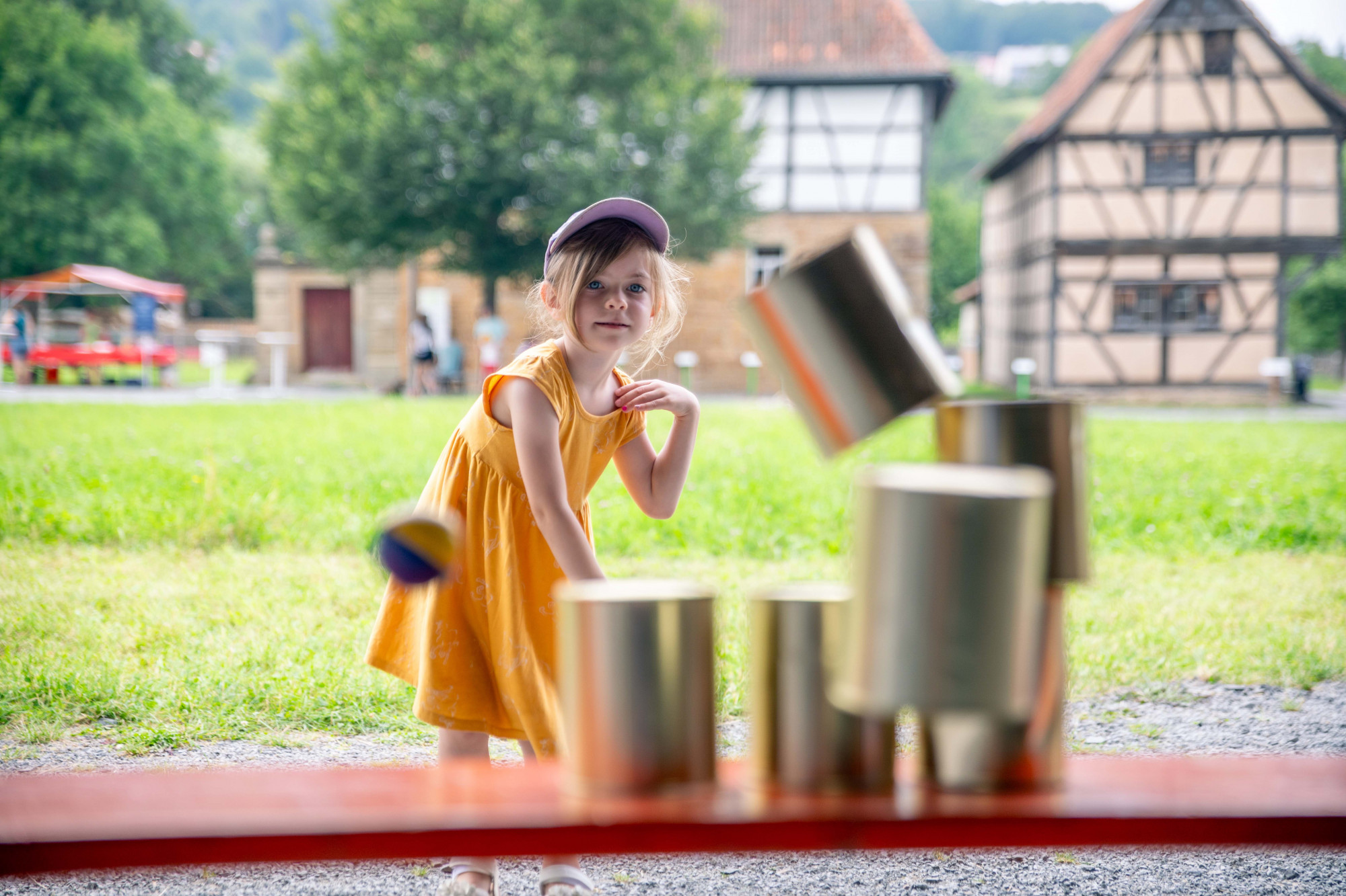 M&auml;dchen beim Dosenwerfen vor einer Wiese mit B&auml;umen und Fachwerkgeb&auml;uden im Hintergrund