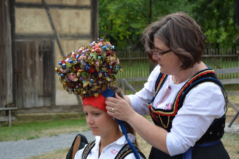Frau in Tracht platziert eine Brautkrone auf dem Kopf einer vor ihr sitzenden Frau