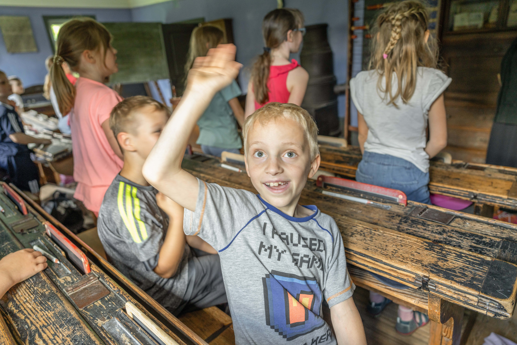 Kind mit grauem T-Shirt hebt in historischem Klassenzimmer die Hand, andere Kinder sitzen an alten Holzb&auml;nken