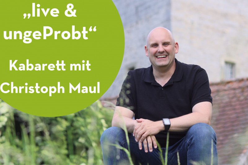 Mann in schwarzem Shirt sitzt vor B&auml;umen und historischem Geb&auml;ude