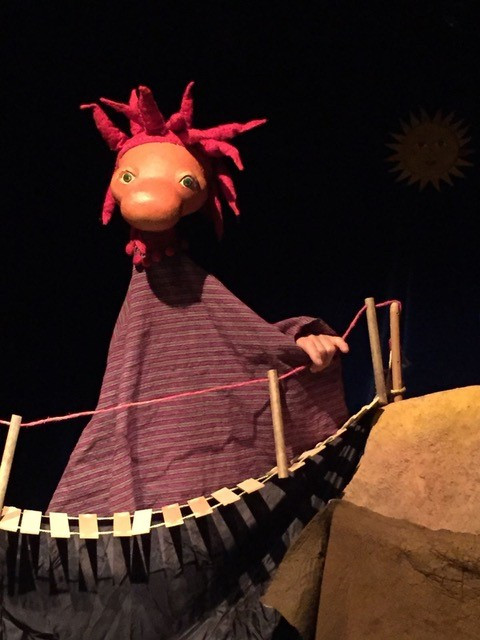 Puppenfigur mit gro&szlig;em Kopf und roten, stacheligen Haaren steht hinter einem kleinen, h&ouml;lzernen H&auml;ngebr&uuml;ckengel&auml;nder auf einer B&uuml;hne des Puppentheaters.