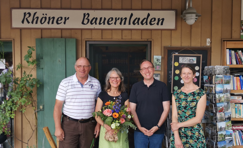 Vier Personen stehen vor dem Eingang des "Rh&ouml;ner Bauernladen", eine Frau h&auml;lt einen Blumenstrau&szlig;, alle l&auml;cheln.