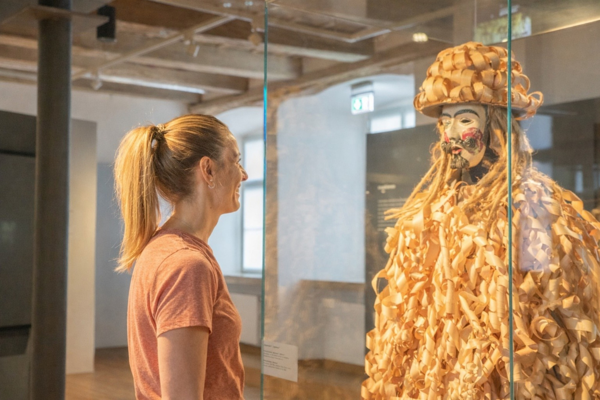 Frau betrachtet Fastnachtskost&uuml;m mit Holzmaske und Pappelsp&auml;nen in Vitrine