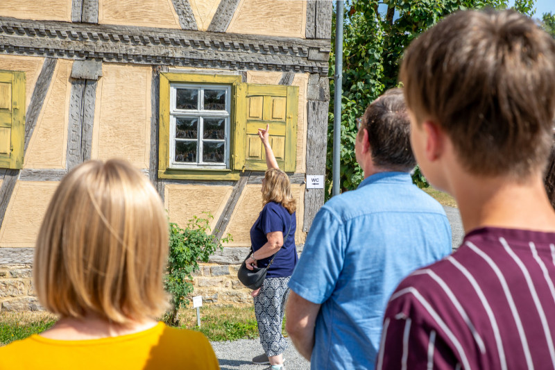 Frau zeigt einer Gruppe von Menschen vor einem Fachwerkhaus mit gr&uuml;nen Fensterl&auml;den etwas an der Fassade