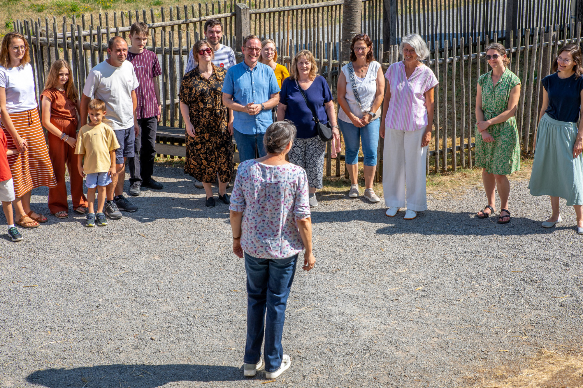 Gruppe von Menschen steht im Freien vor einem Holzzaun und h&ouml;rt einer Frau mit kurz geschnittenem Haar und gemustertem Oberteil zu, die ihnen den R&uuml;cken zugewandt ist
