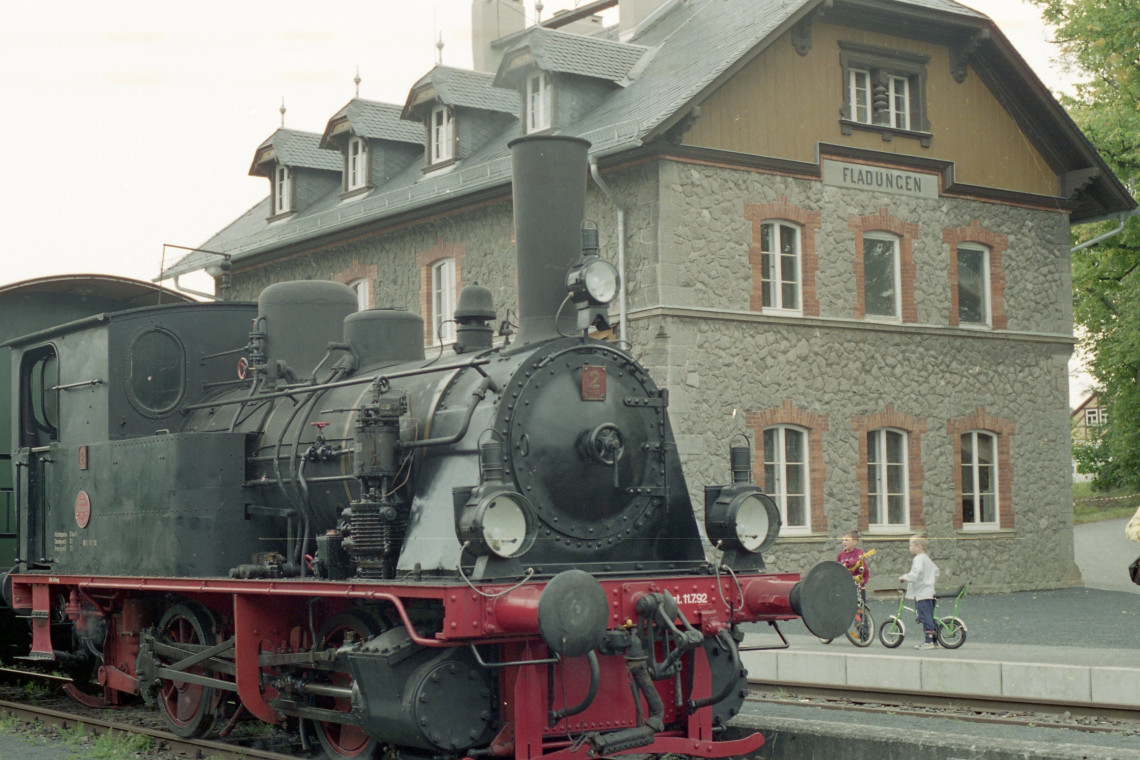 Dampflok vor Bahnhofsgebäude mit der Aufschrift "Fladungen", am Bahnsteig stehen zwei Kindern mit Fahrrädern