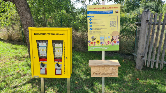 links ein gelber Bienenfutterautomat, rechts ein Hinweisschild, darunter eine Holzbox mit der Aufschrift "Kapselr&uuml;ckgabe" 