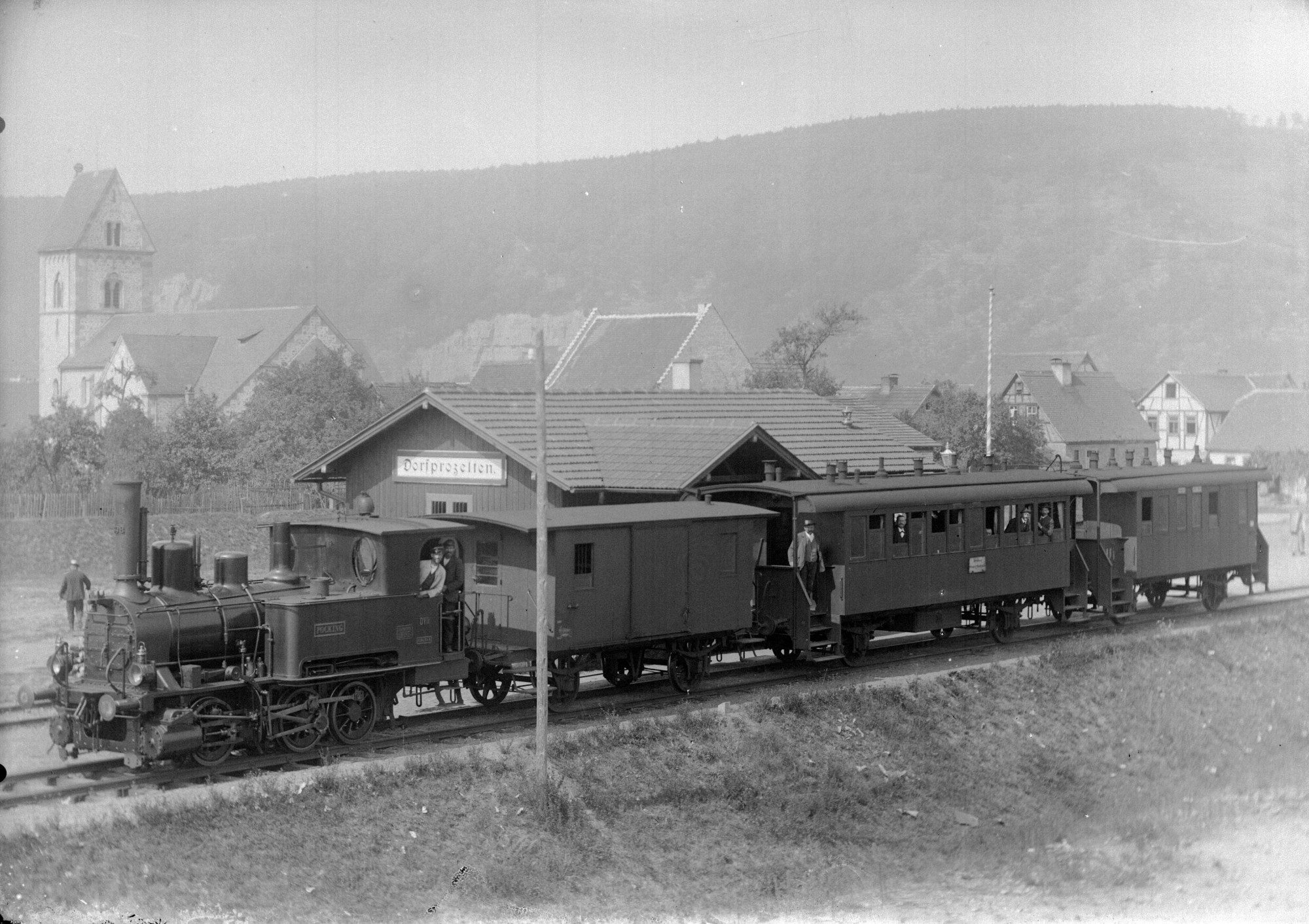 Schwarz-wei&szlig;e Aufnahme einer Dampflokomotive mit mehreren Waggons an einem kleinen Bahnhof vor einer h&uuml;geligen Landschaft