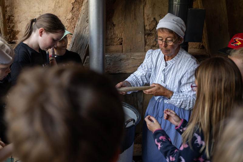 Ältere Frau in historischer Kleidung zeigt einer Gruppe von Kindern in einem rustikalen Innenraum eine traditionelle Waschtechnik.