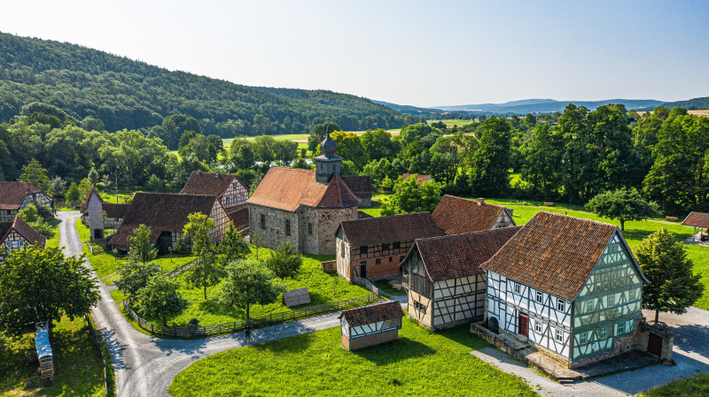 Luftaufnahme eines historischen Freilichtmuseums mit Fachwerkhäusern, einer kleinen Kirche und umgebender grüner Landschaft
