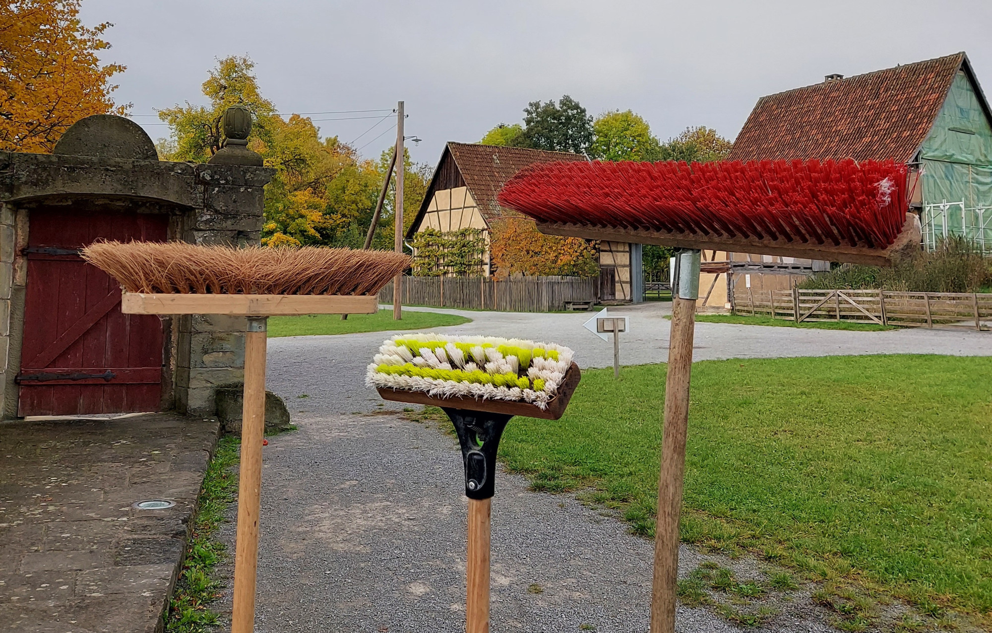 Drei Besen mit Holzstielen stehen auf einem Schotterweg vor einer Wiese und H&auml;usern im Hintergrund