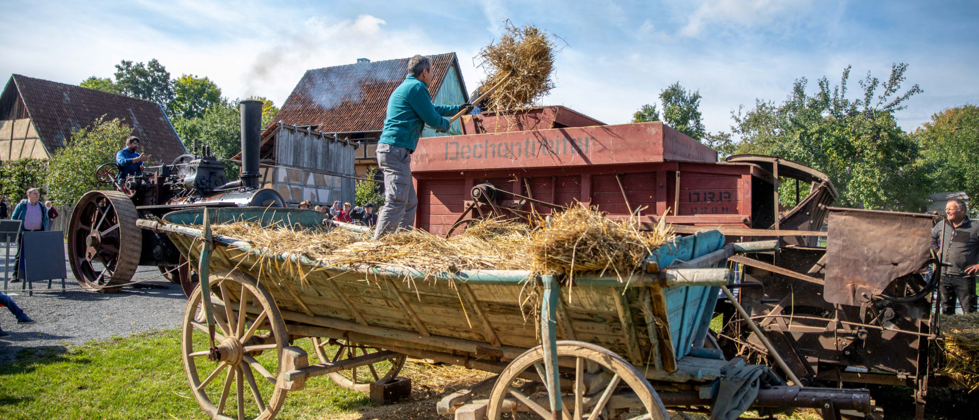 Person wirft Getreide auf eine historische Dreschmaschine im Freilandmuseum neben einem mit Stroh beladenen h&ouml;lzernen Wagen