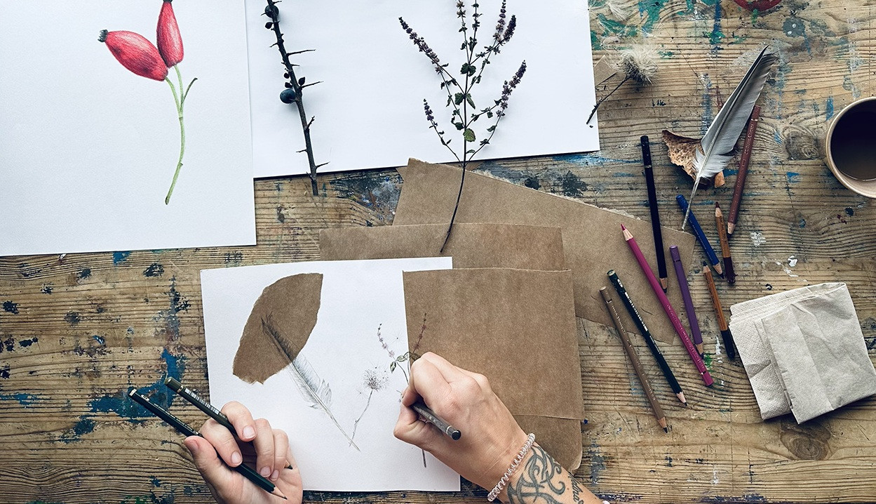Hand zeichnet mit Bleistift eine filigrane Pflanzenskizze auf wei&szlig;em Papier, umgeben von getrockneten Pflanzen, Farbstiften, Pinsel und einer Tasse auf einem Holztisch