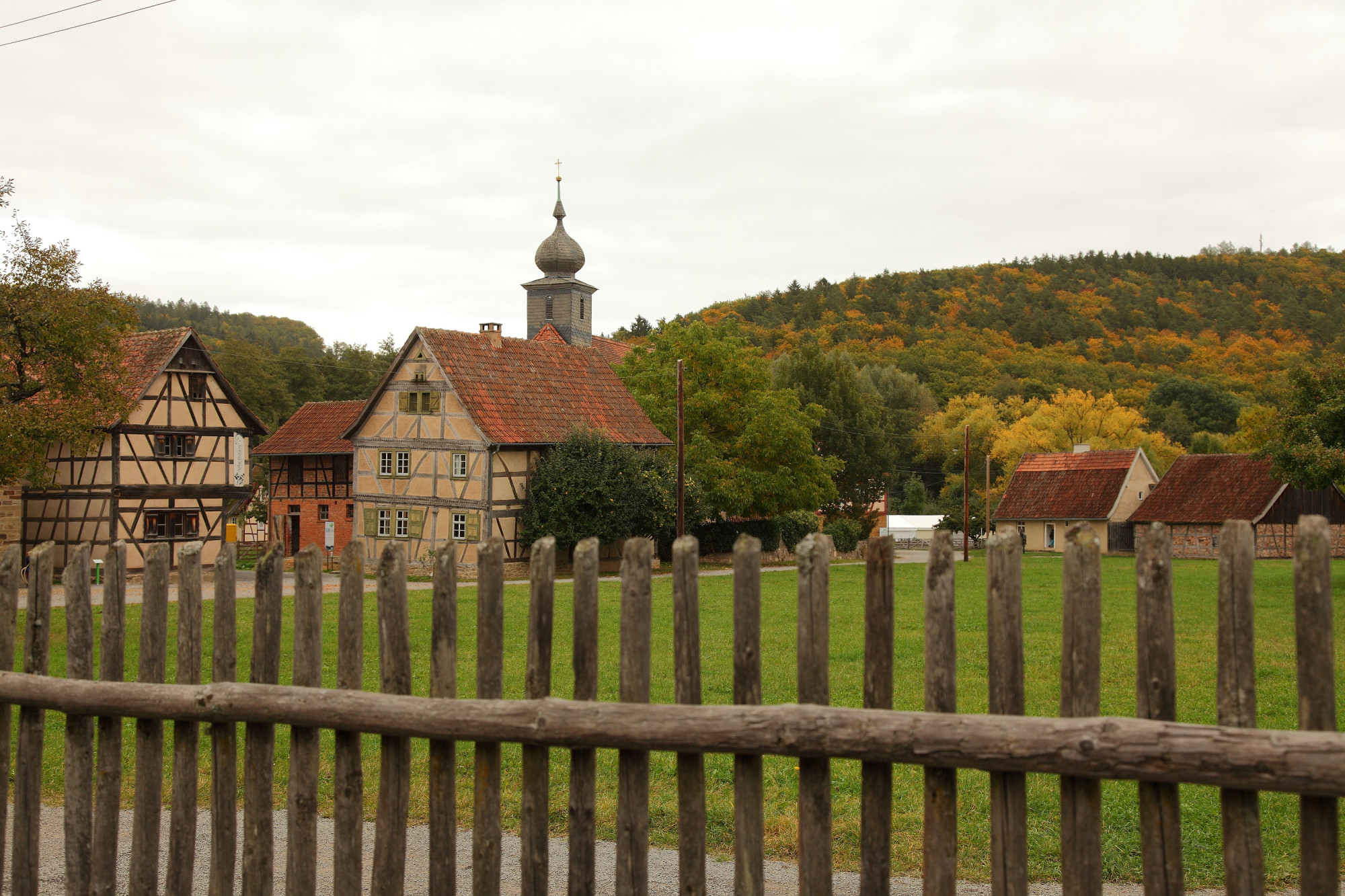 Fachwerkh&auml;user und eine Kirche hinter einem Holzzaun vor herbstlich gef&auml;rbtem Wald