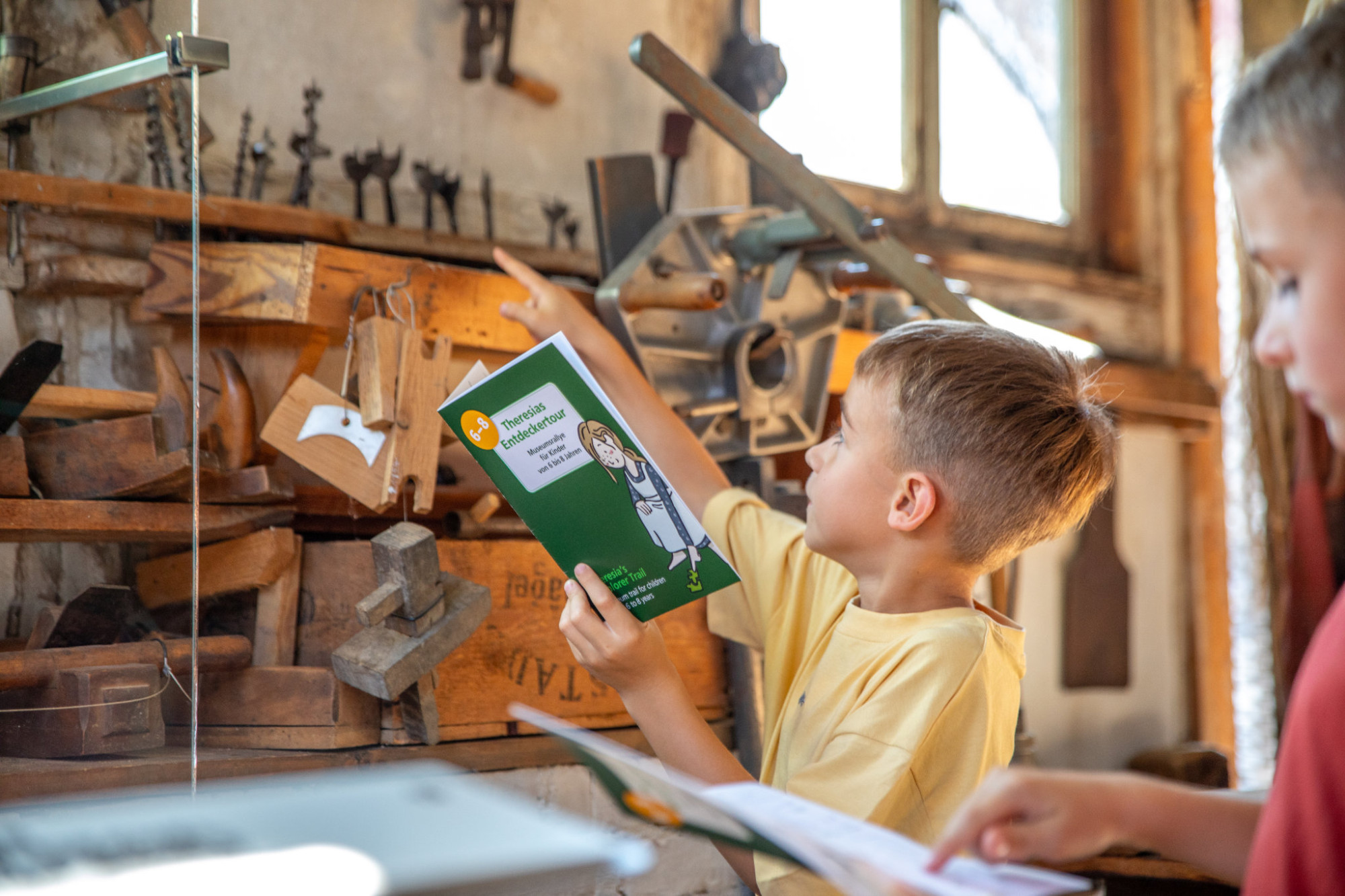 Junge in gelbem T-Shirt h&auml;lt ein gr&uuml;nes Rallyheft und zeigt auf ein h&ouml;lzernes Modell in einem Museum, im Vordergrund ein weiteres Kind mit Rallyheft